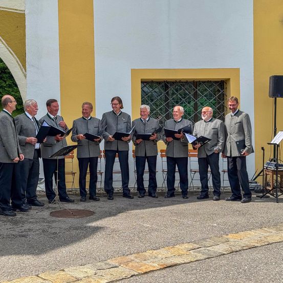 Eine Gruppe von Männern in passenden grauen Anzügen und Krawatten, mit Notenblättern in der Hand, steht vor einem Gebäude mit einem Lautsprecher und einem Fenster.