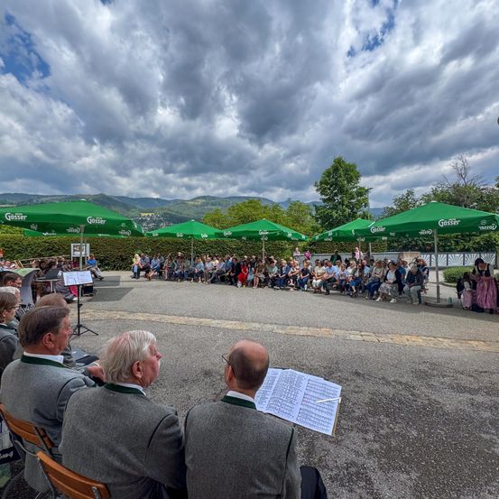 Eine Gruppe von Menschen in traditioneller Kleidung sitzt in einem offenen Bereich unter grünen Schirmen, wahrscheinlich bei einem Musikevent. Vorne stehen einige Leute, andere sind sitzend. Die Umgebung ist von viel Grün und Bergen geprägt.