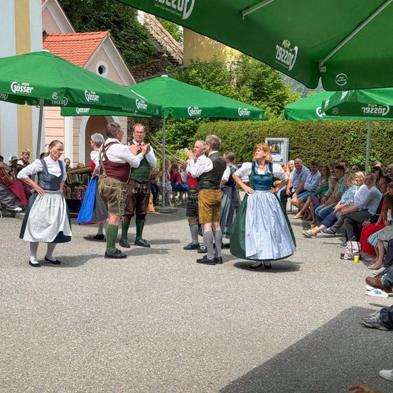 Eine Gruppe von Menschen in traditioneller Kleidung tanzt vor einem Publikum unter grünen Schirmen.