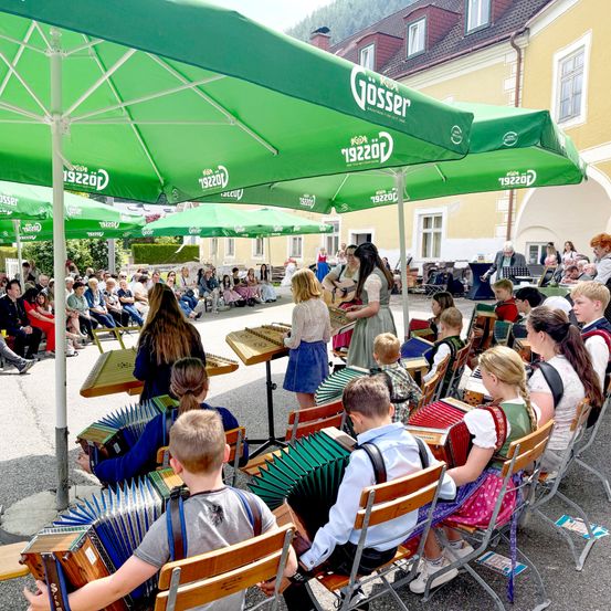 Kinder spielen Akkordeons vor einem Publikum unter grünen Gösser-Markisen. Das Publikum sitzt auf Stühlen und schaut zu.
