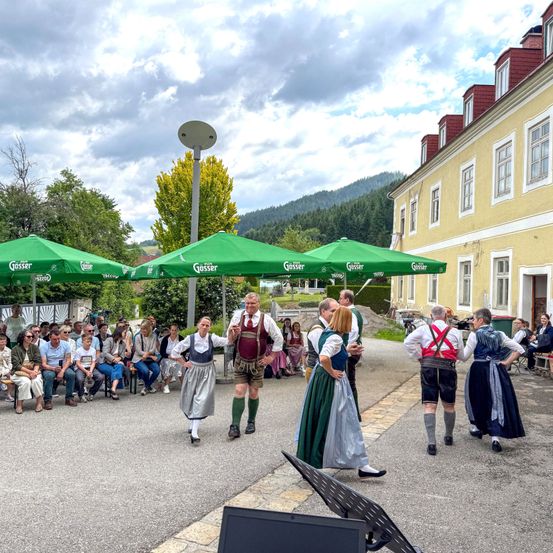 Eine Gruppe von Menschen in traditionellen Kostümen tanzt vor einem gelben Gebäude. Ein Publikum sitzt unter grünen Schirmen. Im Hintergrund befindet sich ein Berg mit Bäumen.