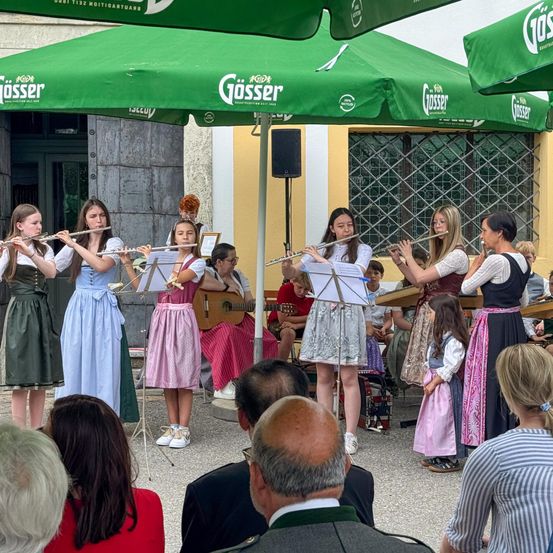 Eine Gruppe junger Frauen in traditioneller Kleidung spielt Flöten unter grünen Schirmen. Dahinter spielen andere Gitarren. Ein Publikum beobachtet sie im Freien.