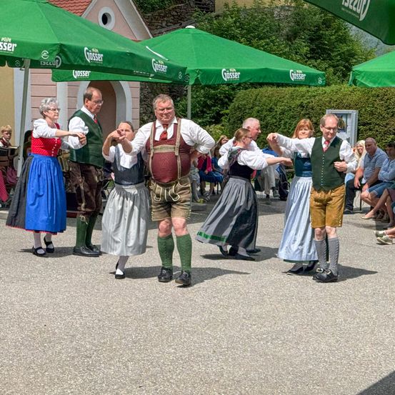 Eine Gruppe von Erwachsenen in traditioneller Kleidung tanzt in einem Freiluftbereich. Sie stehen in einer Reihe mit verbundenen Armen. Hinter ihnen sitzen Leute unter grünen Schirmen. Ein Gebäude befindet sich hinter den Menschen, und ein grüner Busch steht davor.