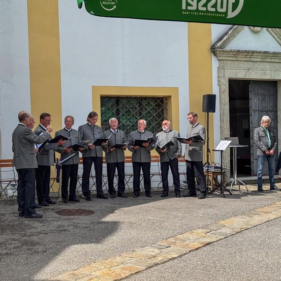 Ein Chor von Männern in passenden grauen Anzügen steht vor einem Gebäude und singt. Ein Mann in einem grünen Jackett steht vor dem Gebäude.