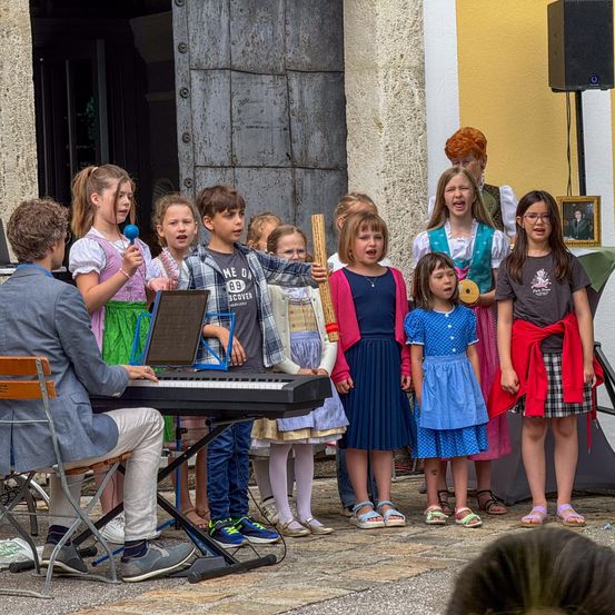 Eine Gruppe von Kindern und ein erwachsener Mann führen vor einem Gebäude auf. Der Mann spielt Klavier, während die Kinder singen.