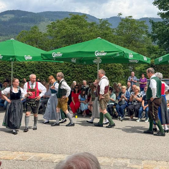 Eine Gruppe von Männern und Frauen in traditioneller deutscher Kleidung führt einen Tanz vor einem Publikum auf. Die Umgebung besteht aus grünen Schirmen, einem bergigen Hintergrund und sitzenden Zuschauern unter einem bewölkten Himmel.