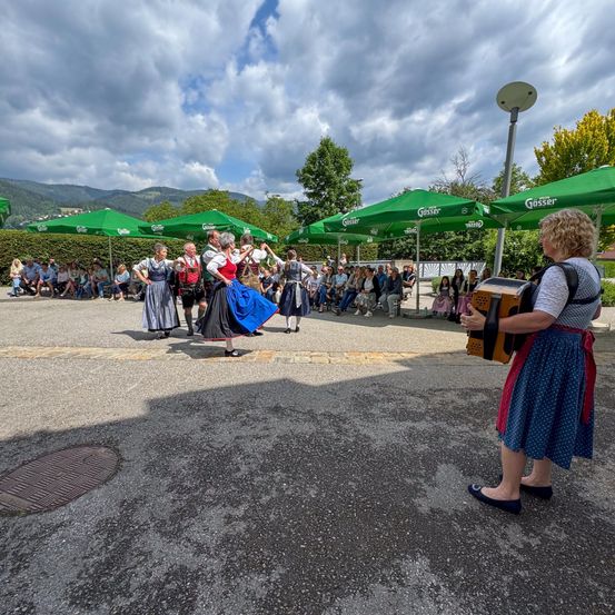 Eine Gruppe von Menschen in traditioneller Kleidung tanzt unter grünen Schirmen, während ein Publikum zusieht. Eine Frau spielt in der Nähe eine Akkordeon.