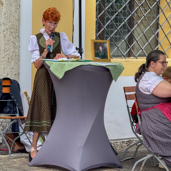 Eine ältere Frau in einem gepunkteten Kleid spricht in ein Mikrofon an einem Tisch. Ein gerahmtes Porträt und ein grüner Stoff sind auf dem Tisch. Eine Frau in einem gepunkteten Kleid sitzt neben ihr.