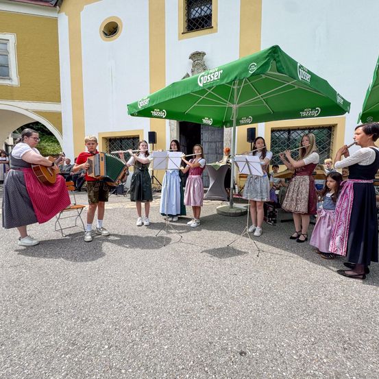 Eine Gruppe von Kindern und Erwachsenen spielt Musik unter einem grünen Schirm auf einem Innenhof. Sie spielen Instrumente wie Flöten und Akkordeons.