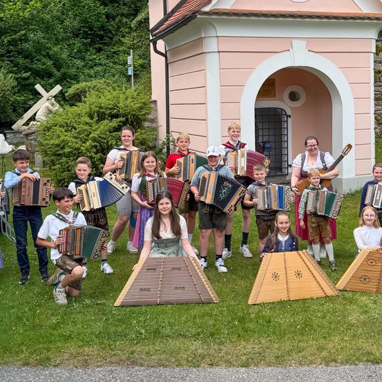 Eine Gruppe junger Musiker spielt Akkordeons vor einem rosa Gebäude. Sie knien und sitzen auf dem Gras. Dahinter befindet sich ein Gebäude mit Fenster und Rundbogentür.