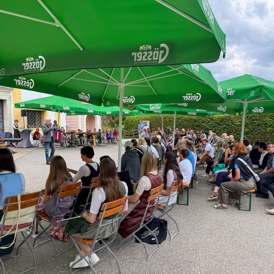 Eine Menschenmenge versammelt sich in einem Außenbereich unter grünen Sonnenschirmen. Ein Mann steht vorne, möglicherweise hält er eine Rede. Viele Menschen sitzen, während andere stehen und zuschauen.