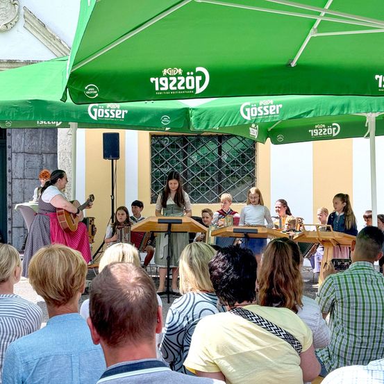 Eine Gruppe von Kindern spielt Instrumente unter grünen Gosser-Bäumen vor einem Publikum. Eine Frau steht links und spielt Gitarre.