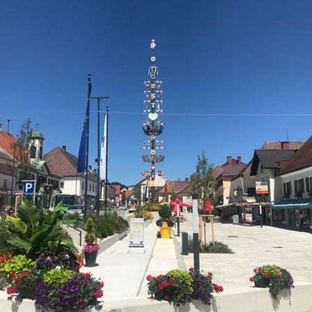 Ein Stadtplatz mit dekorativen Straßenlaternen, Gebäuden und einem klaren Himmel. Verschiedene Pflanzen und Blumen schmücken den Bereich, mit Flaggen und einem Parkplatzschild sichtbar.