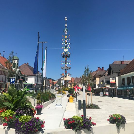 Ein Stadtplatz mit dekorativen Straßenlaternen, Gebäuden und einem klaren Himmel. Verschiedene Pflanzen und Blumen schmücken den Bereich, mit Flaggen und einem Parkplatzschild sichtbar.