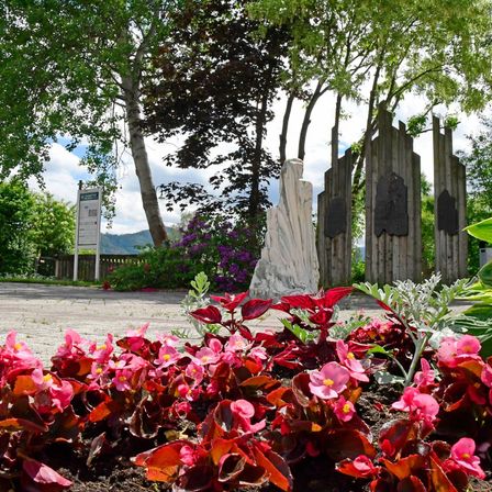 Ein Park mit leuchtend rosa und roten Blumen, einer Statue und hölzernen Strukturen. Im Hintergrund befinden sich Bäume und Grünflächen.