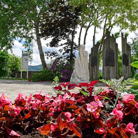 Ein Park mit leuchtend rosa und roten Blumen, einer Statue und hölzernen Strukturen. Im Hintergrund befinden sich Bäume und Grünflächen.