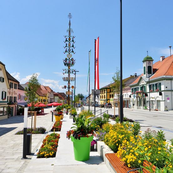 Eine Straße in einer Stadt mit einem geschmückten Turm, Flaggen, Gebäuden und bunten Blumen. Die Straße ist gesäumt von Bäumen, Pflanzen und Straßenlaternen.
