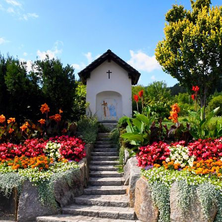 Eine kleine weiße Kapelle mit einem Kreuz auf der Vorderseite ist von bunten Blumen umgeben. Steinstufen führen zum Eingang, und ein Baum mit gelben Blüten steht in der Nähe.
