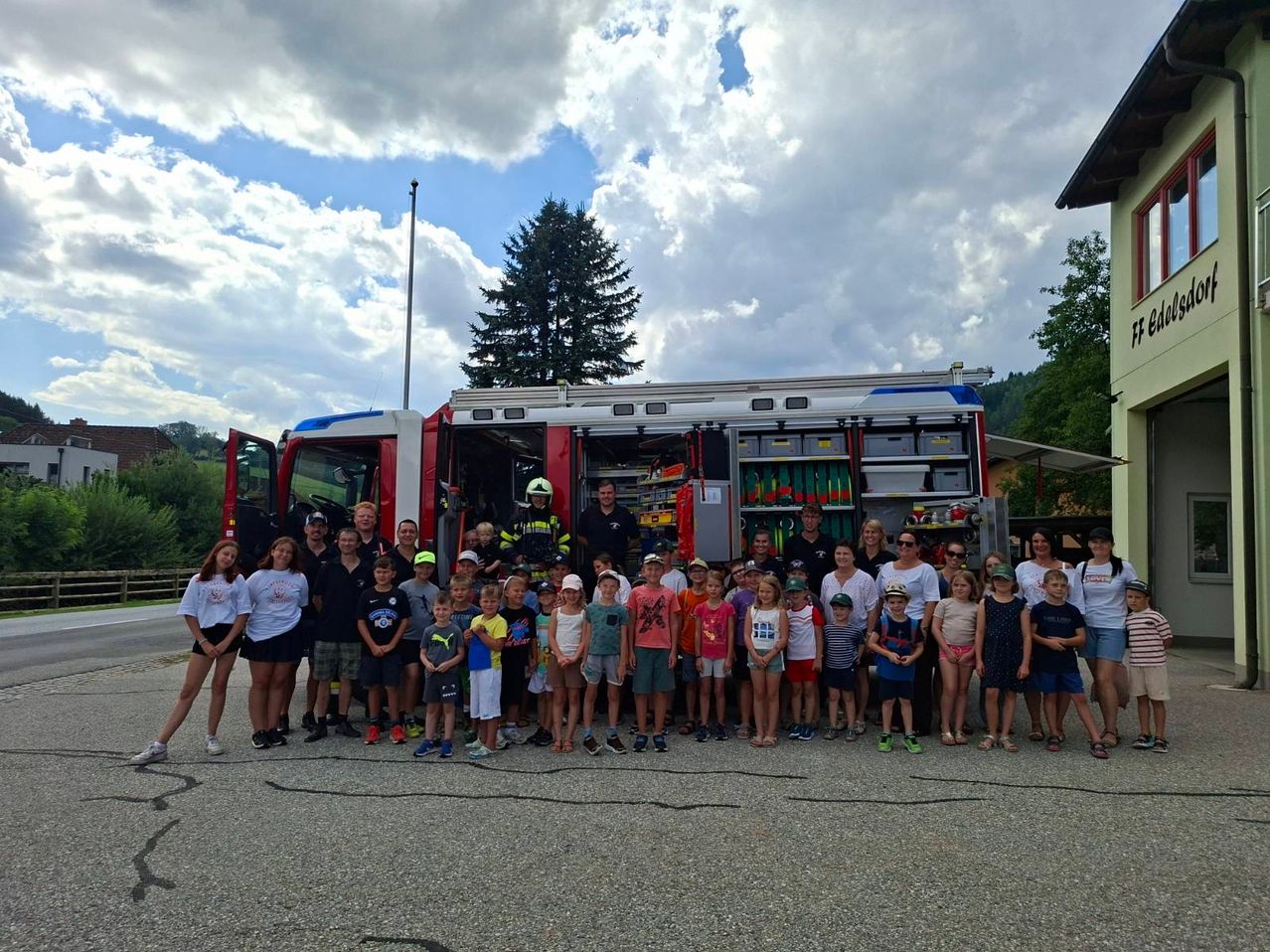 Eine Gruppe von Kindern und Erwachsenen posiert für ein Foto vor einem Feuerwehrauto an einem sonnigen Tag.