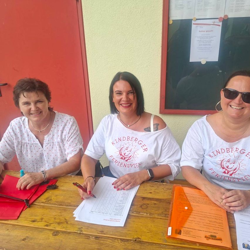 Drei Frauen sitzen an einem Holztisch und lächeln. Die Frau in der Mitte hält einen Stift und Papier. Sie tragen weiße T-Shirts mit rotem Text. Ein roter Tür und ein Bulletin Board sind im Hintergrund.
