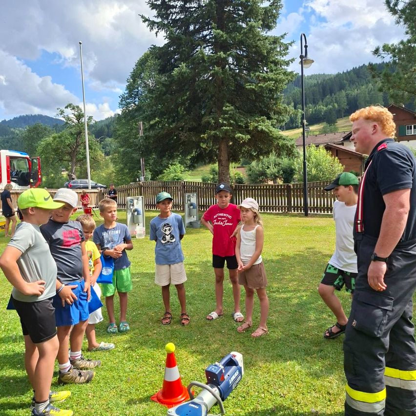 Eine Gruppe von Kindern und ein Feuerwehrmann stehen auf einem Rasenfeld mit einem Feuerlöscher und einem Verkehrskegel. Die Kinder tragen Hüte und Turnschuhe. Hinter ihnen steht ein Holzzaun, ein Baum und ein Haus. In der Ferne gibt es Berge und einen bewölkten Himmel.