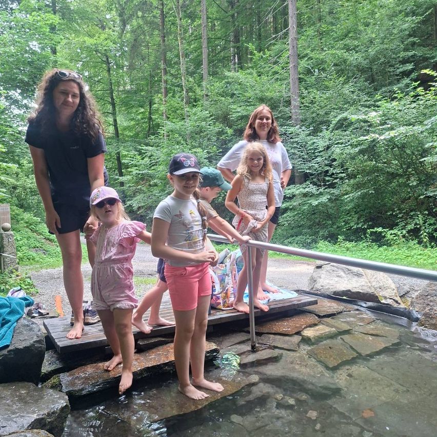 Eine Familie mit fünf Personen steht auf einer Holzplattform über einer natürlichen Quelle, mit zwei jungen Mädchen in rosa Kleidern und Hüten, einem Jungen in einer Kappe und zwei Frauen, eine mit Sonnenbrille auf dem Kopf.