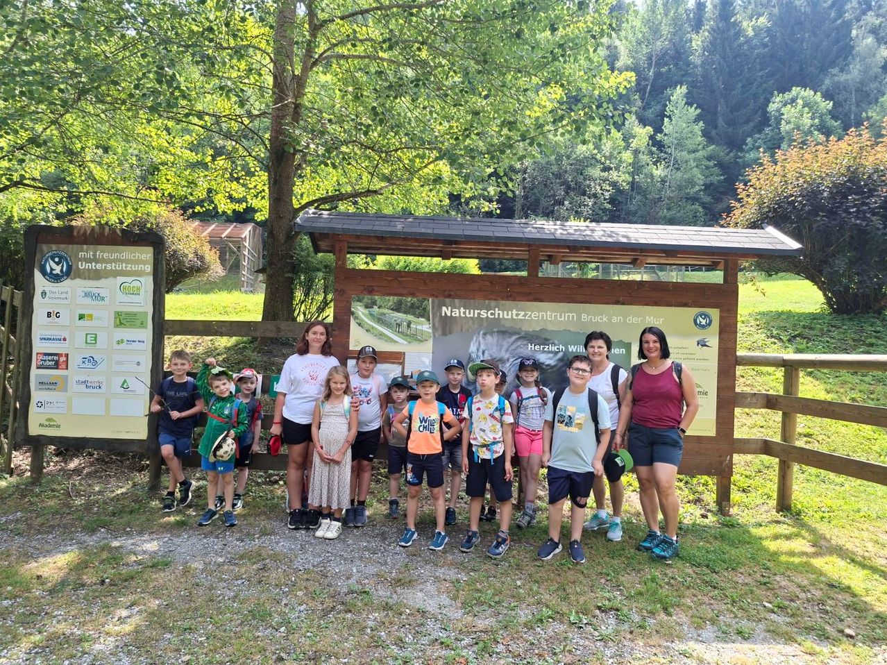 Eine Gruppe von Kindern und Erwachsenen posiert für ein Foto vor einem hölzernen Schild im Park. Das Schild hat ein Bild eines Bären und einige Texte.