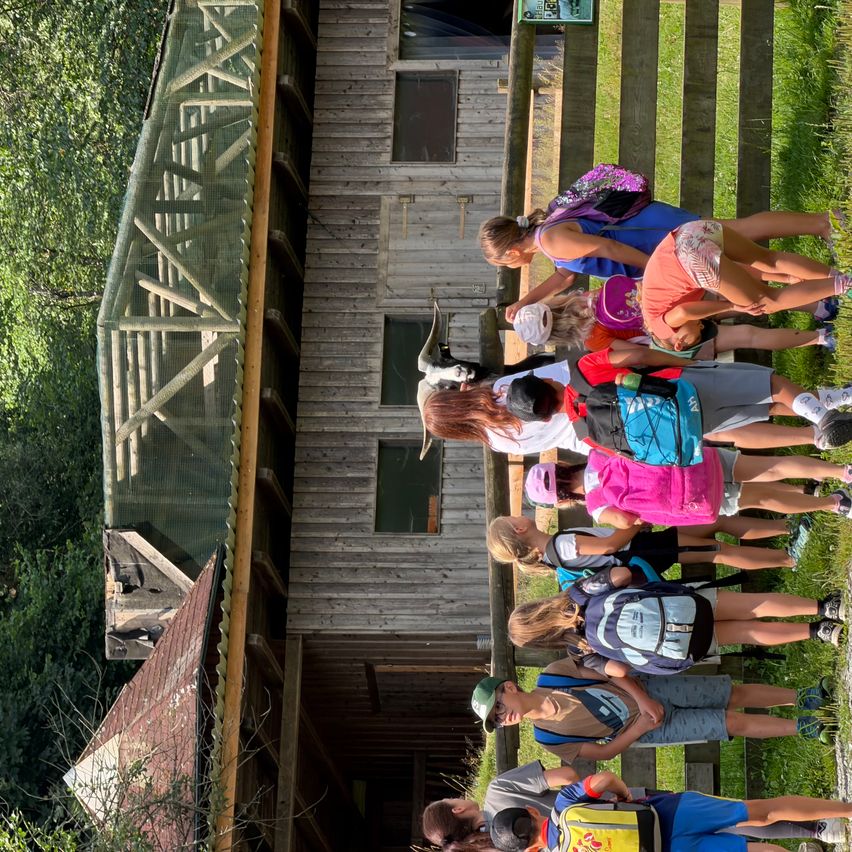 Eine Gruppe von Kindern mit Rucksäcken und Turnschuhen steht in einer Reihe vor einem Holzgebäude.