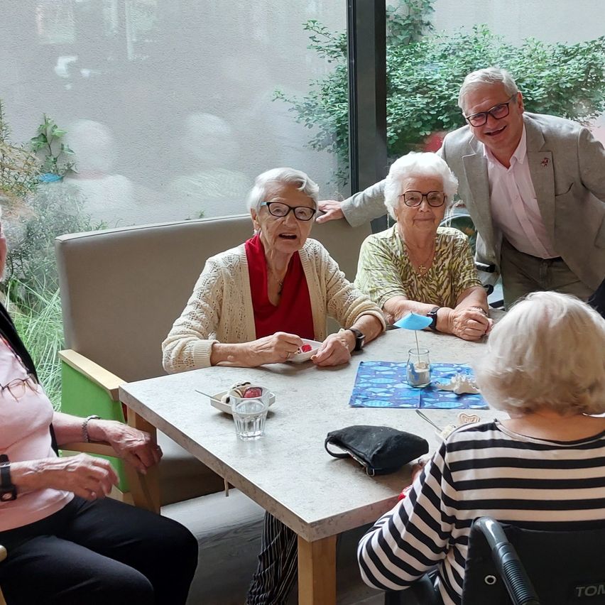 Eine Gruppe älterer Frauen und ein Mann sitzen um einen Tisch und lächeln und interagieren. Auf dem Tisch stehen ein Becher, ein Glas und ein kleiner Teller. Der Hintergrund zeigt eine Glaswand mit Pflanzen.