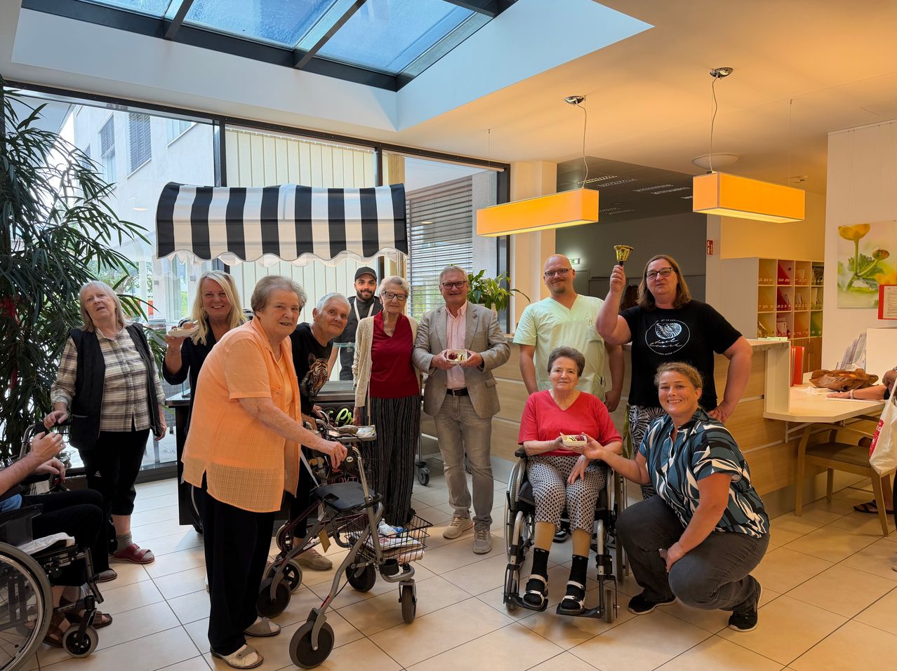 Eine Gruppe von Menschen, darunter einige im Rollstuhl, versammelt sich in einer Lobby, lächelt und posiert für ein Foto. Eine Frau im Rollstuhl hält eine kleine Glocke.