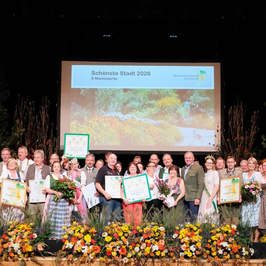 A group of people stand on a stage holding certificates and flowers, with a large screen behind them showing a landscape and the text 'Schönste Stadt 2026 9 Nominierte'.
