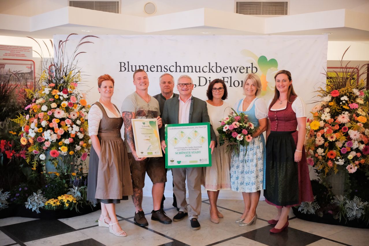 A group of people in traditional German attire stand in front of a banner with the text 'Blumenschmuckbewerb, Die Flor'. They are holding certificates and flowers. The man in the middle is wearing glasses and a suit.