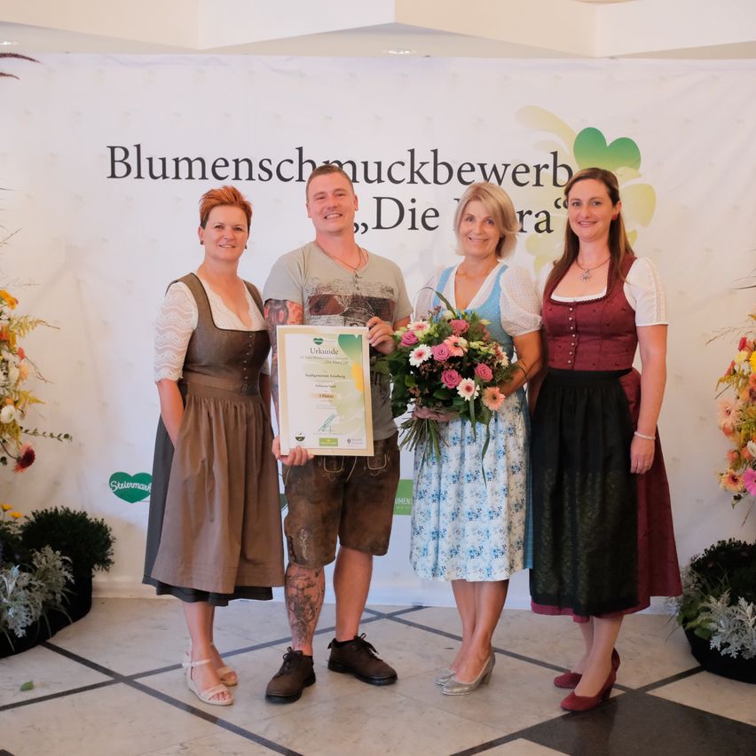 Four people stand in front of a white backdrop with the words 'Blumenschmuckbewerb'. The man in the middle holds a certificate and a bouquet of flowers. The women wear traditional clothing.