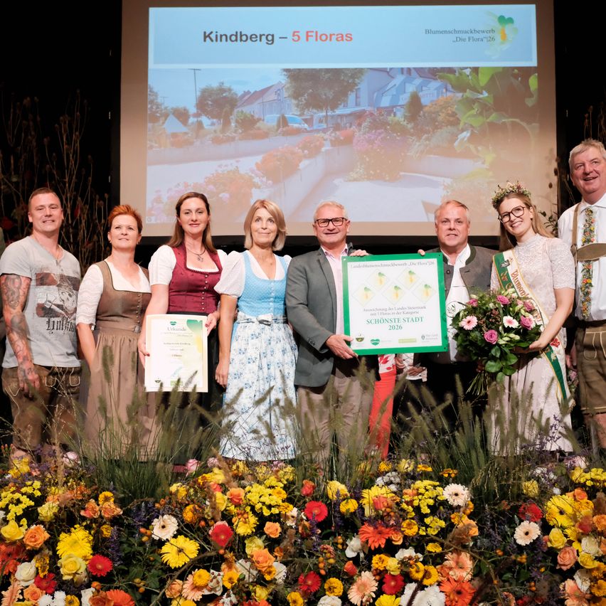 A group of people stand on stage holding certificates and flowers, with a projector screen behind them displaying an image of a house and garden.
