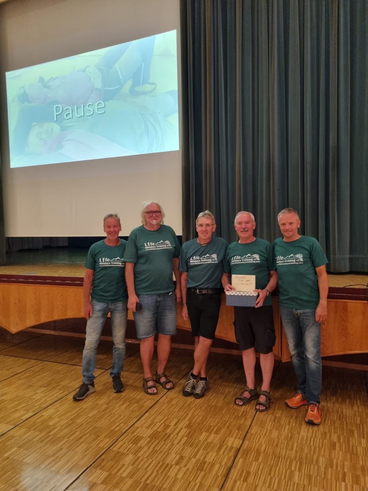 Five men in green shirts stand on a stage, one holding a box, with a projector screen behind them displaying the word 'Pause'.