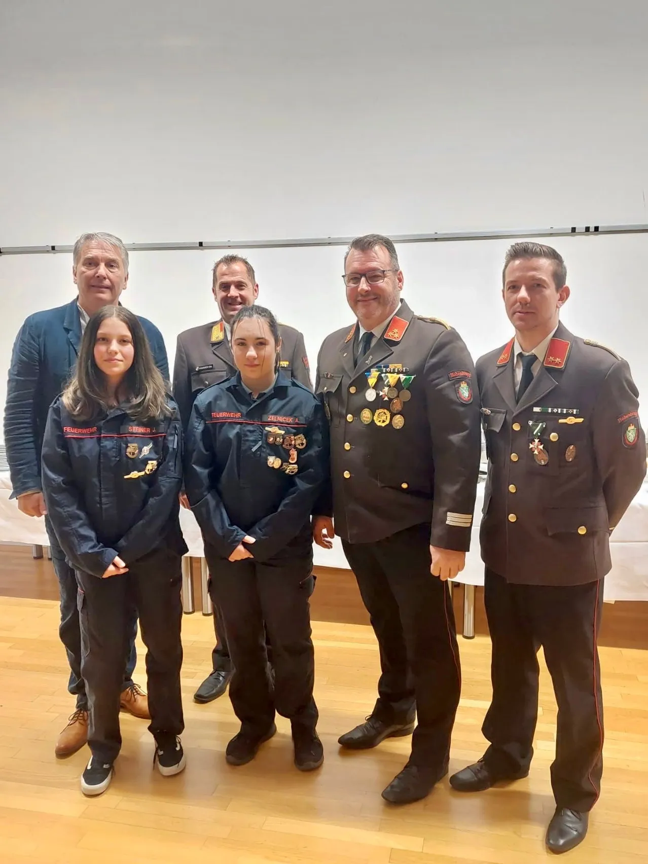 Fünf Personen in Uniformen stehen in einem Raum. Zwei junge Frauen tragen blaue Jacken mit rotem Saum. Hinter ihnen stehen drei Männer in Militäruniformen, alle mit Medaillen. Sie posieren für ein Foto.