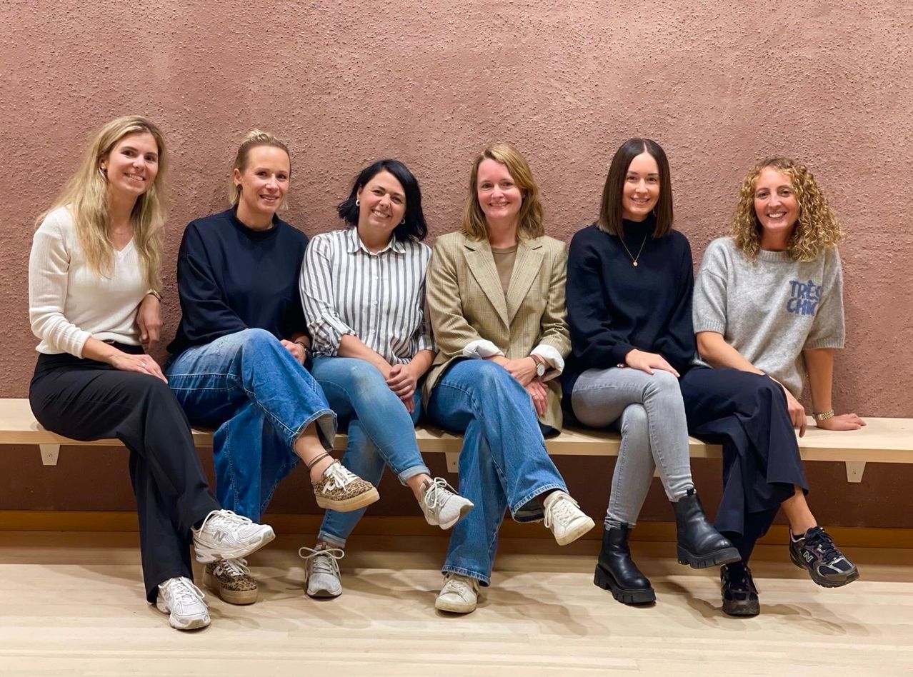 Eine Gruppe von Frauen sitzt auf einer Bank, lächelt und posiert für ein Foto. Sie sind lässig gekleidet, mit verschiedenen Stilen und Farben. Die Wand hinter ihnen ist rosa.