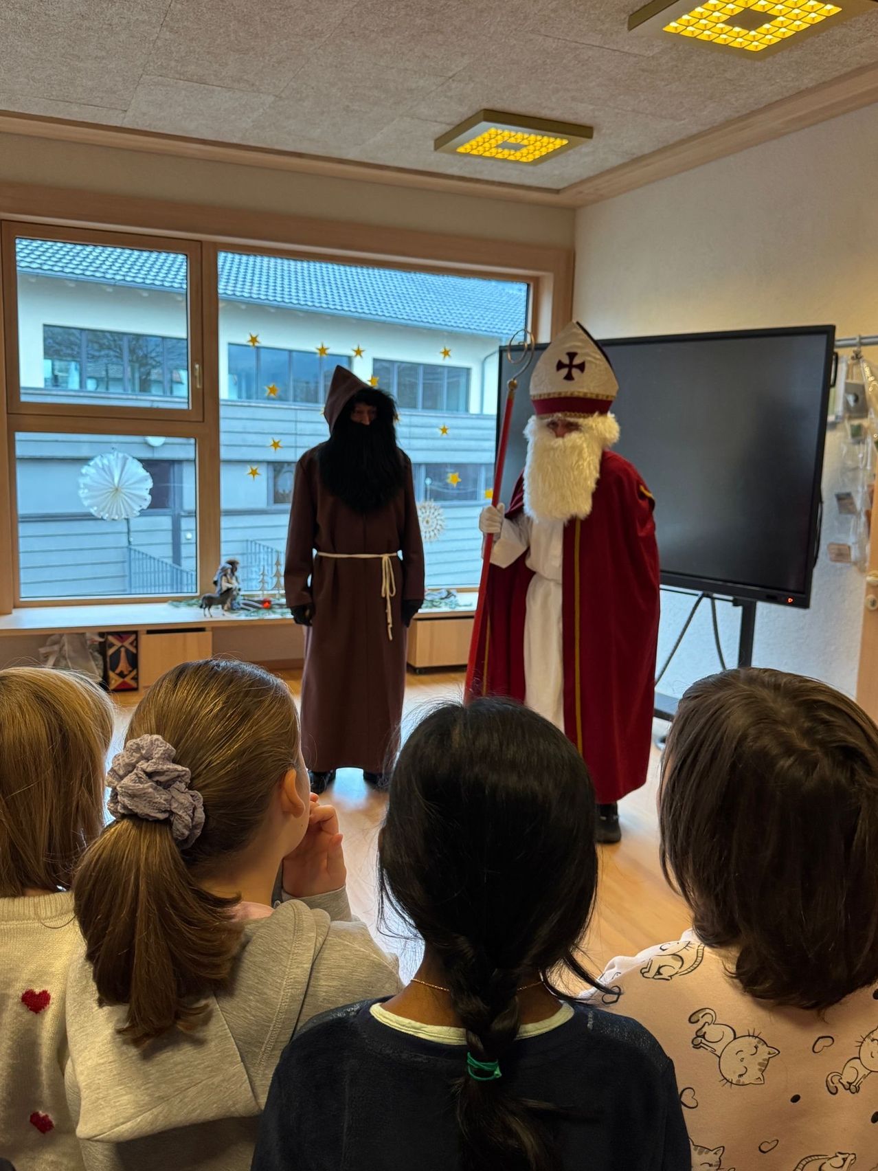 Zwei Personen, gekleidet als St. Nikolaus und ein Mönch, stehen in einem Raum mit Kindern. Ein Fernseher und ein Fenster mit Blick auf ein Gebäude sind im Hintergrund.