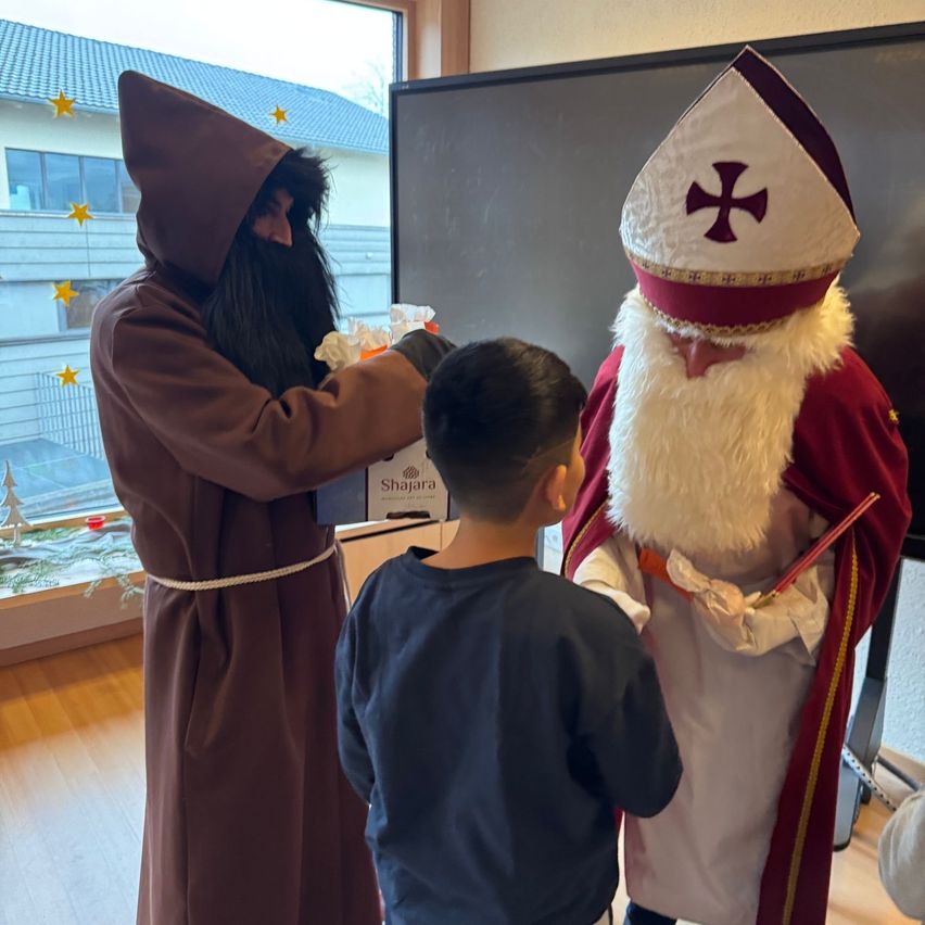 Ein Mann in einem braunen Gewand reicht einem Jungen ein verpacktes Geschenk, während eine Figur als Heiliger Nikolaus in der Nähe steht. Sie befinden sich in einem weihnachtlich geschmückten Raum.