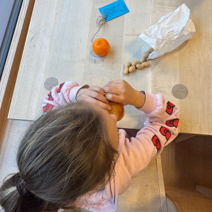 Ein Kind in rosa Pyjamas sitzt am Tisch und isst ein Sandwich, mit einer Orange und Erdnüssen in der Nähe. Ein blauer Papieranhänger befindet sich auf dem Tisch.