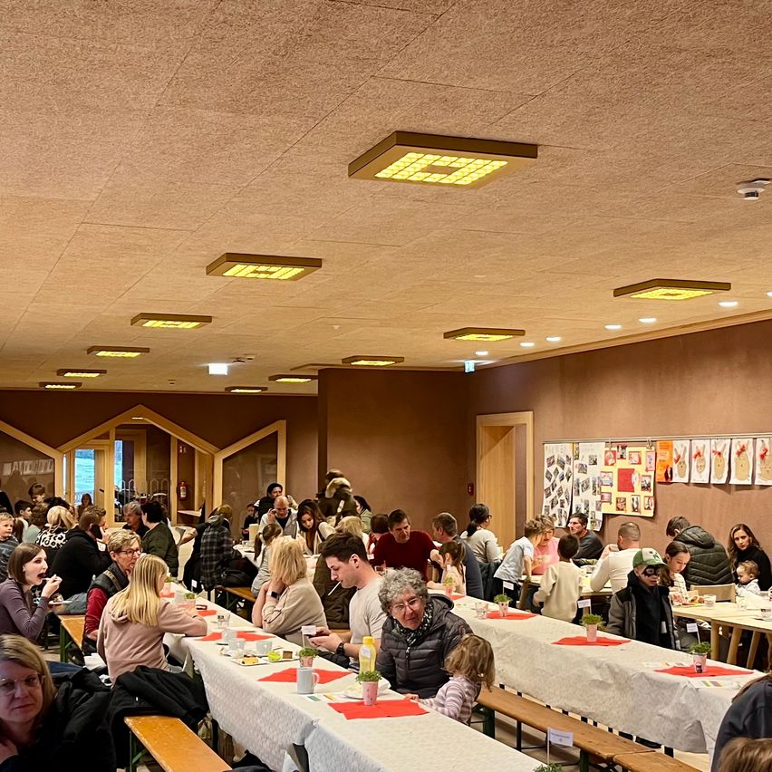 Eine große Gruppe von Menschen sitzt an Tischen in einem Raum mit weißen Tischdecken und Topfpflanzen. Die Wände sind mit Kunstwerken geschmückt. Der Raum hat eine Holzdecke mit mehreren Lichtern.