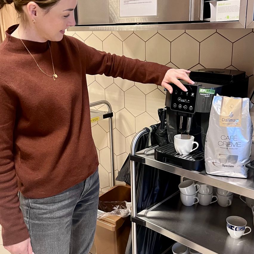 Eine Frau bereitet Kaffee mit einer Maschine zu, mit einer Tüte Cafe Creme daneben, vor einer gefliesten Wand stehend.