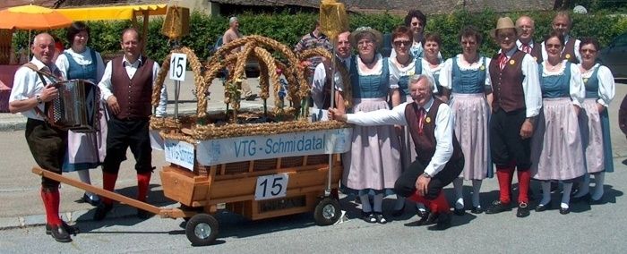 Eine Gruppe von Menschen in traditioneller bayerischer Kleidung steht neben einem geschmückten Holzwagen, wobei einer ein Akkordeon spielt und ein anderer kniet. Der Wagen trägt die Nummer 15 und steht 'VTG-Schmidala'.
