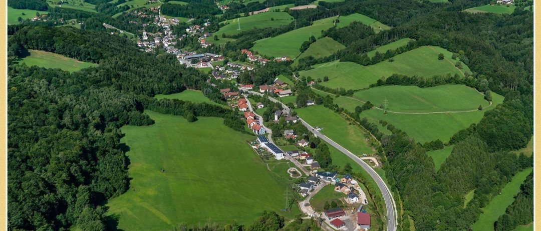 Eine Luftaufnahme einer ländlichen Landschaft mit grünen Feldern, Häusern und einer Straße, umgeben von Bäumen und Bergen unter einem klaren Himmel.