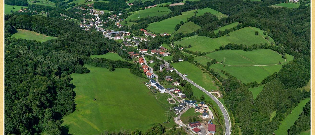 Eine Luftaufnahme einer ländlichen Landschaft mit grünen Feldern, Häusern und einer Straße, umgeben von Bäumen und Bergen unter einem klaren Himmel.