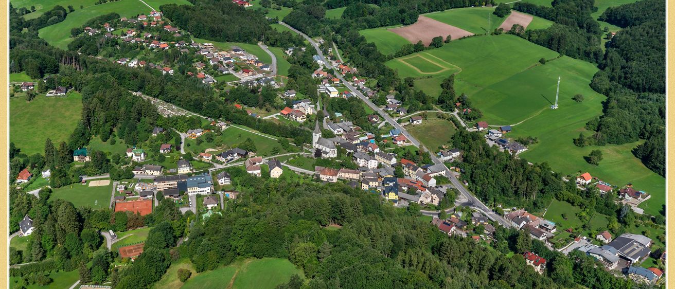 Luftaufnahme eines Dorfes, umgeben von üppigen grünen Feldern und dichten Wäldern, mit einem klaren blauen Himmel darüber.