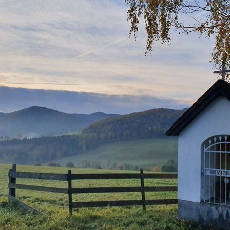 Eine kleine Kapelle mit einem Kreuz neben einem Holzzaun, mit Bergen und Nebel im Hintergrund.
