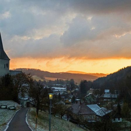 Sonnenuntergang über einer kleinen Stadt mit einem Kirchturm, schneebedecktem Boden und einem Pfad, der durch das Dorf führt.