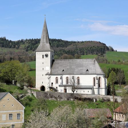 Eine weiße Kirche mit einem Turm und einer Uhr befindet sich auf einem Hügel, umgeben von grünen Feldern und Bäumen. Sie hat ein graues Dach, gewölbte Fenster und einen kleinen Vorbau.