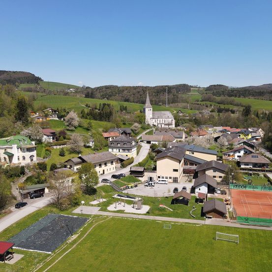 Luftaufnahme eines ländlichen Ortes mit Kirche, Häusern, Tennisplätzen und einem grünen Feld unter blauem Himmel.